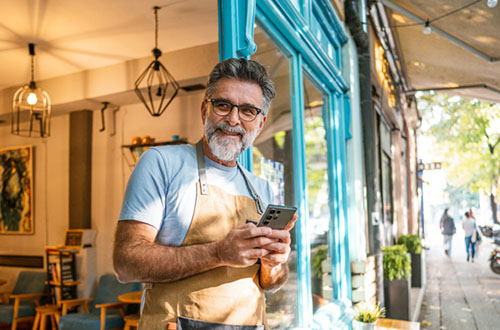 Mature man owner with an apron using phone in front of the café